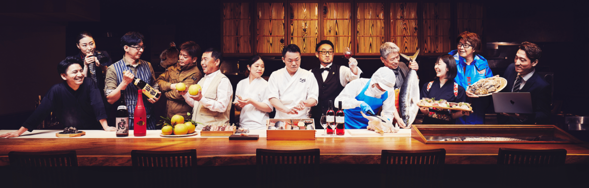 13人の食のプロが並んでいる写真。生産者、料理人、バーテンダーなど、それぞれの持場をイメージする服装やポーズを、長いカウンターの中でとっている。カメラ目線の人は一人もいない。