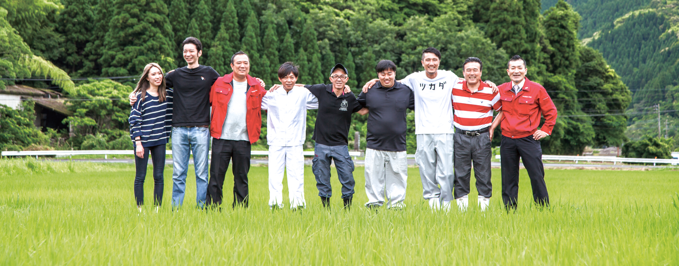 緑の深い山を背景に並んで立つ契約農家の皆さん。9人が肩を組み笑顔でカメラに目線を向け写真におさまっている。左から3人目の方が来ている白いシャツには、カタカナで「ツカダ」の文字が書かれている。