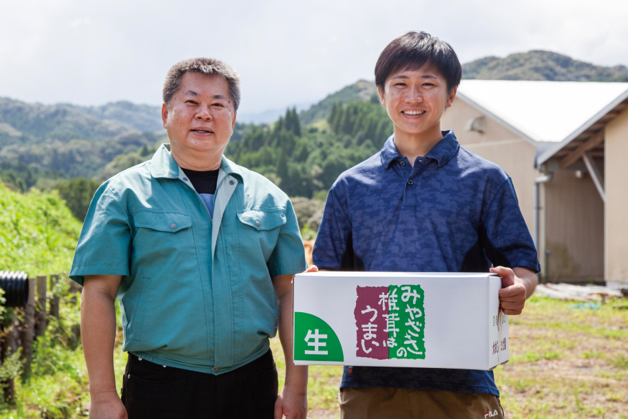 契約農家のお二人。向かって右に立つ方は白い段ボールを両手で抱えており、その側面には「みやざきの椎茸はうまい」の文字が印刷されている。