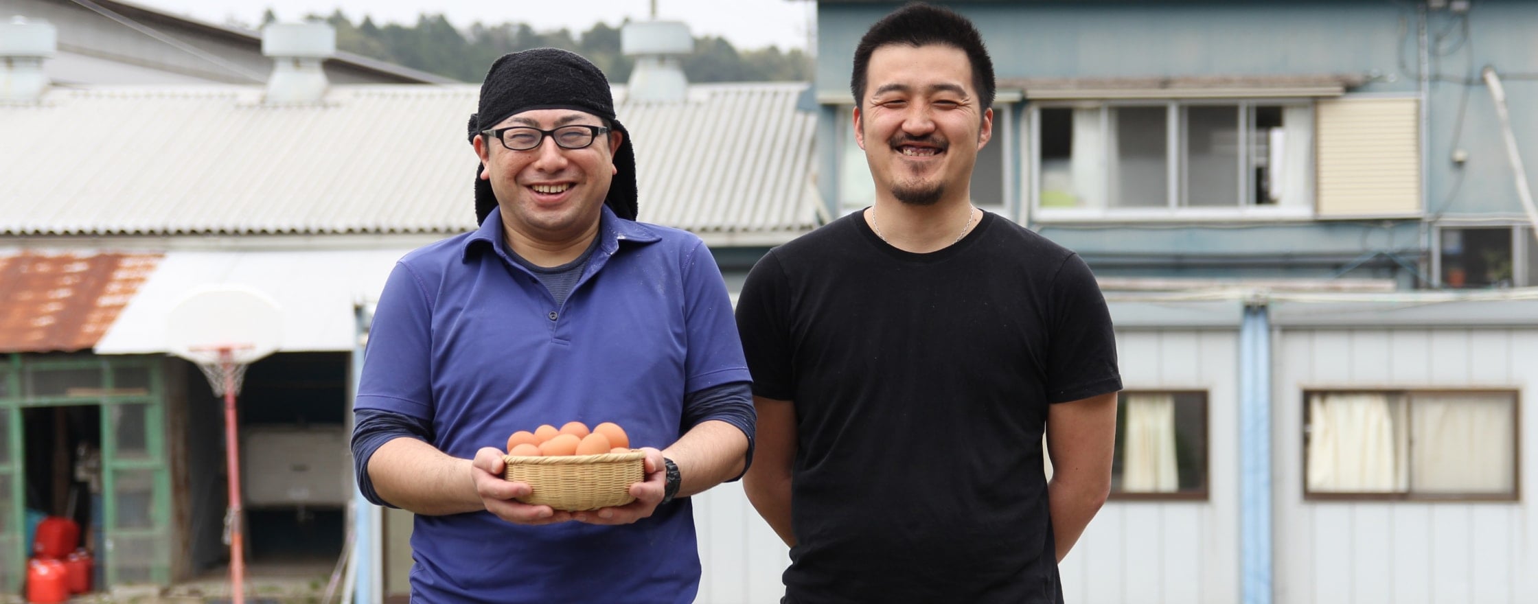 飼育場を背景に正面を向いて笑顔で並んでいる生産者のお二人。向かって左の方は、茶色の卵がいっぱい入った竹のカゴを両手で持っている