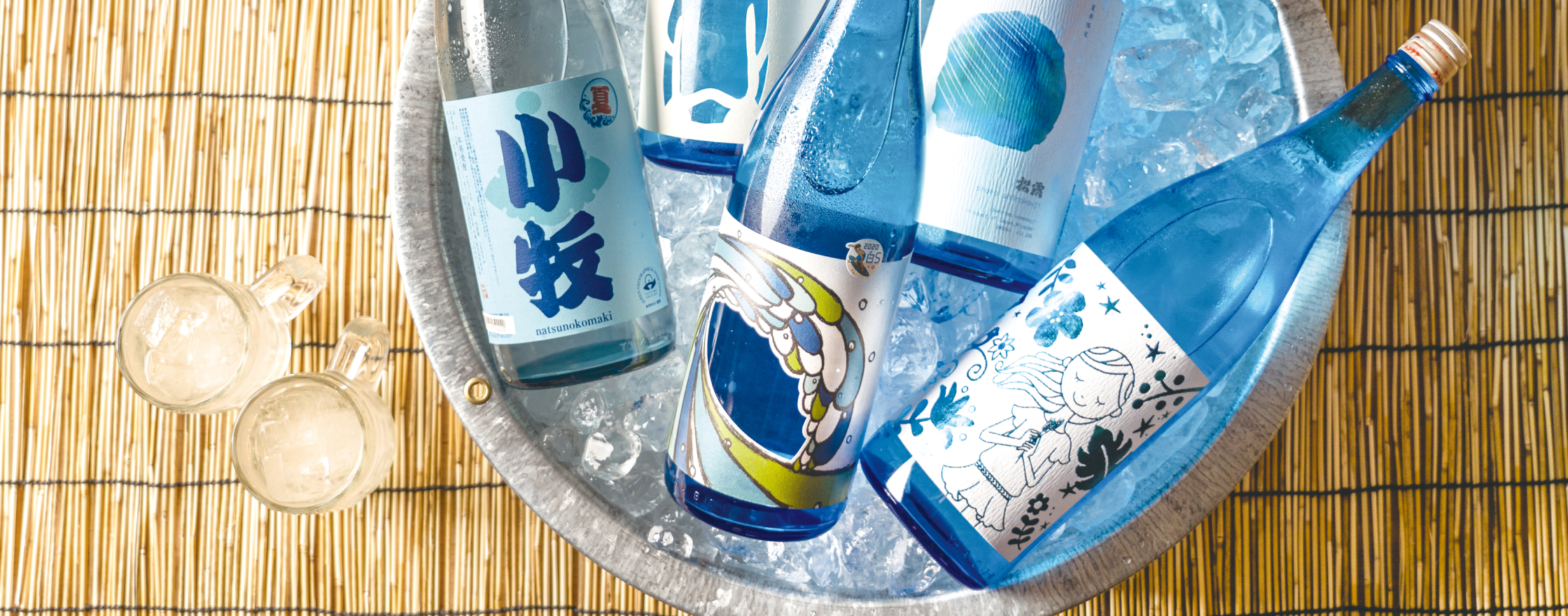 氷がはられたタライで冷やされている5本の焼酎。清涼感のある青い瓶と青系のデザインラベル。タライの横には氷の入ったジョッキが２つ、下には竹すだれがしいてある涼しげな写真
