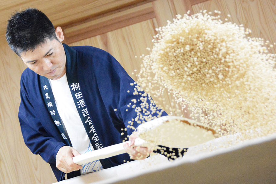 白い麹を大きなヘラでかき混ぜる職人。紺色の法被で真剣な表情。