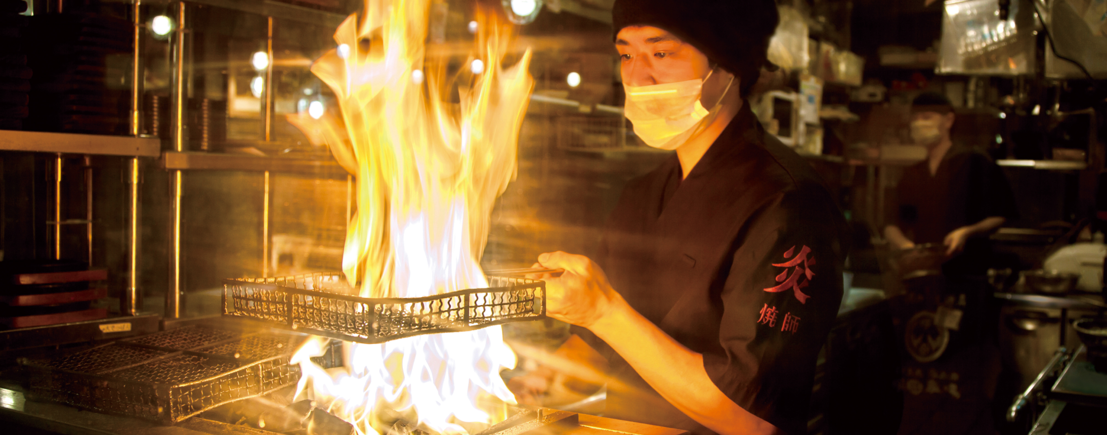 調理場で大きな炎を前にして鶏を焼く職人。右手で四角い網カゴを持ち、背丈と同じくらいの炎が網を貫いている。職人が来ている黒い服の右腕には「炎焼師」の赤い刺繍が見える